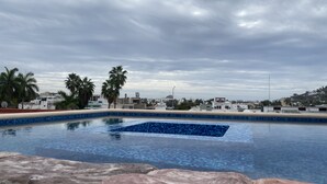 A swimming pool with a tiled edge, surrounded by rocks and palm trees, with buildings in the background under a cloudy sky. - Hotel Tierra Marina Centro Histórico (Mazatlán)