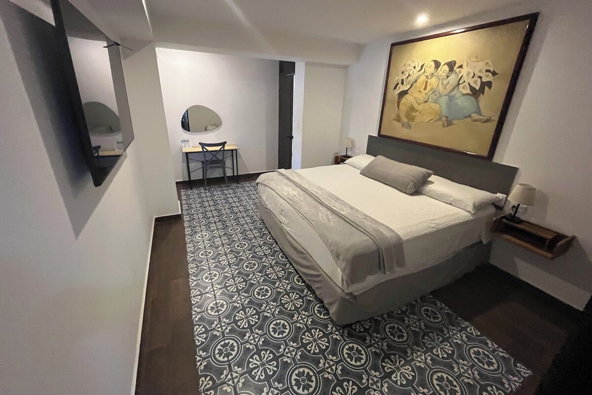 Una habitación de hotel moderna con una cama grande, un escritorio, una silla, una mesita, una mesita de noche y un cuadro en la pared.