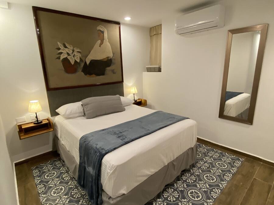 A bedroom with a large bed, a nightstand with a lamp, a wall-mounted air conditioner, and a large mirror.