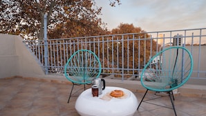 Una terraza con dos sillas turquesas, un reposapiés blanco, una cafetera de metal y un croissant sobre el reposapiés.