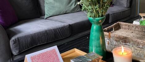 A cozy living room with a grey sofa, a green vase with white flowers, a tray with a card and a candle, and a painting on the wall.