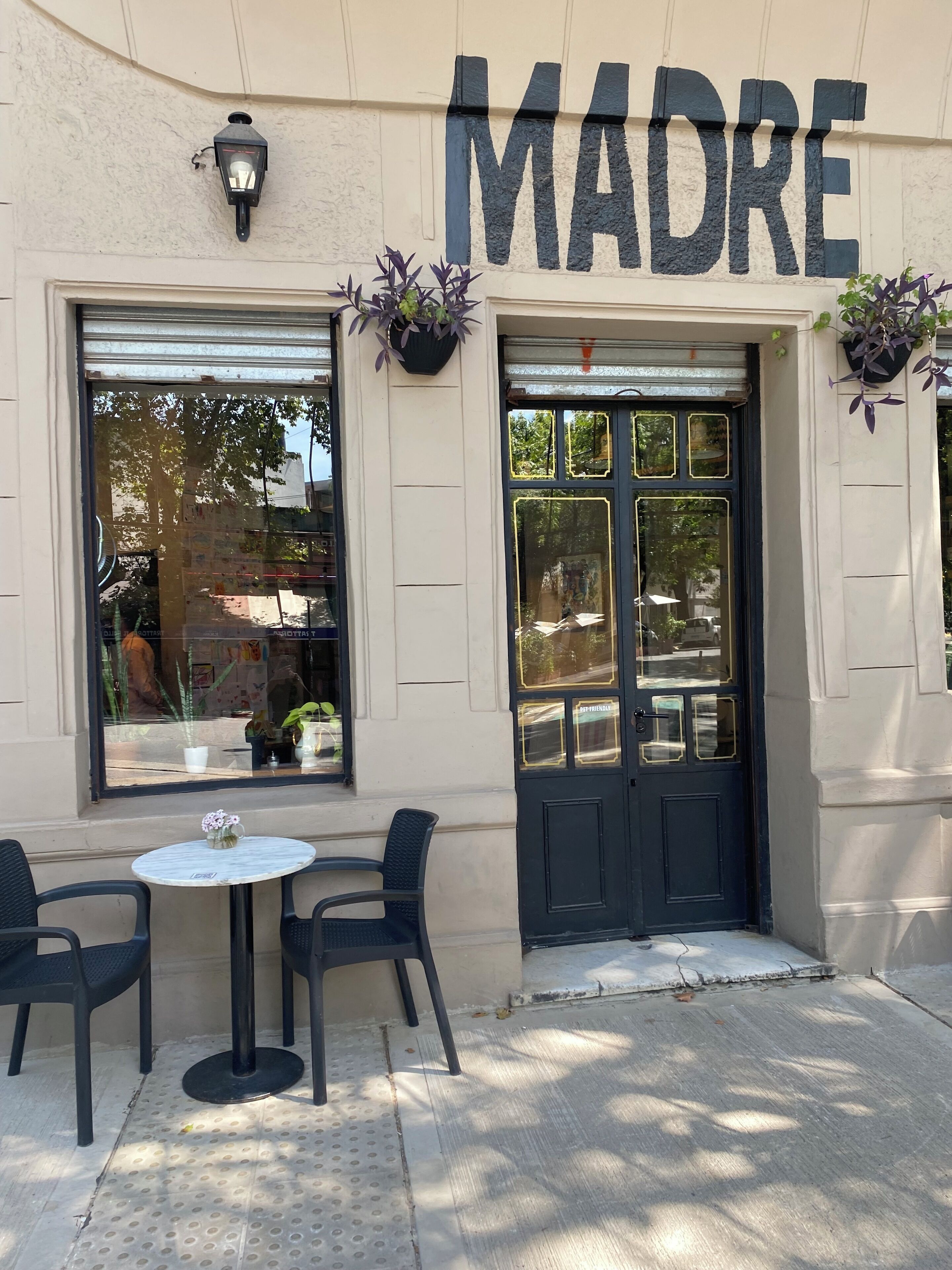 A building with the sign 'MADRE' above the entrance. There are two outdoor tables with chairs, and potted plants are placed on either side of the entrance.