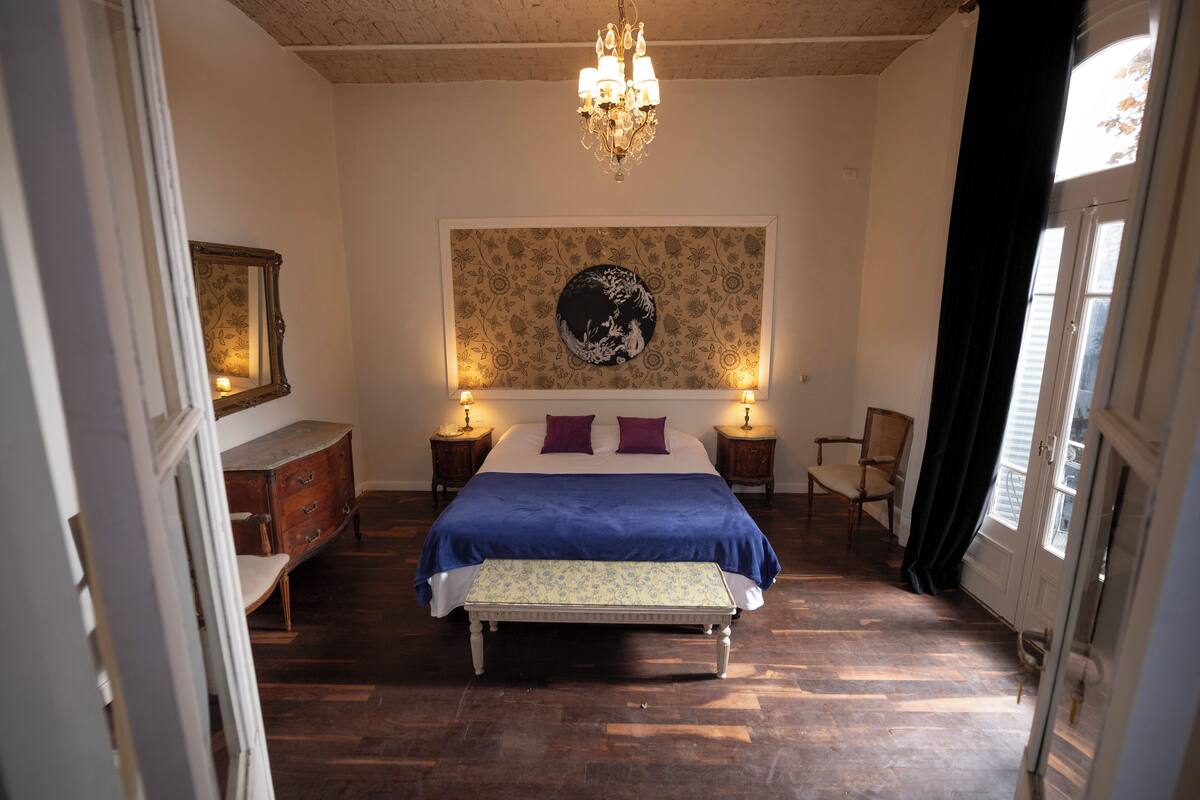 A bedroom with a large bed, a bench, a dresser, a chair, a chandelier, and a wall with a circular design.