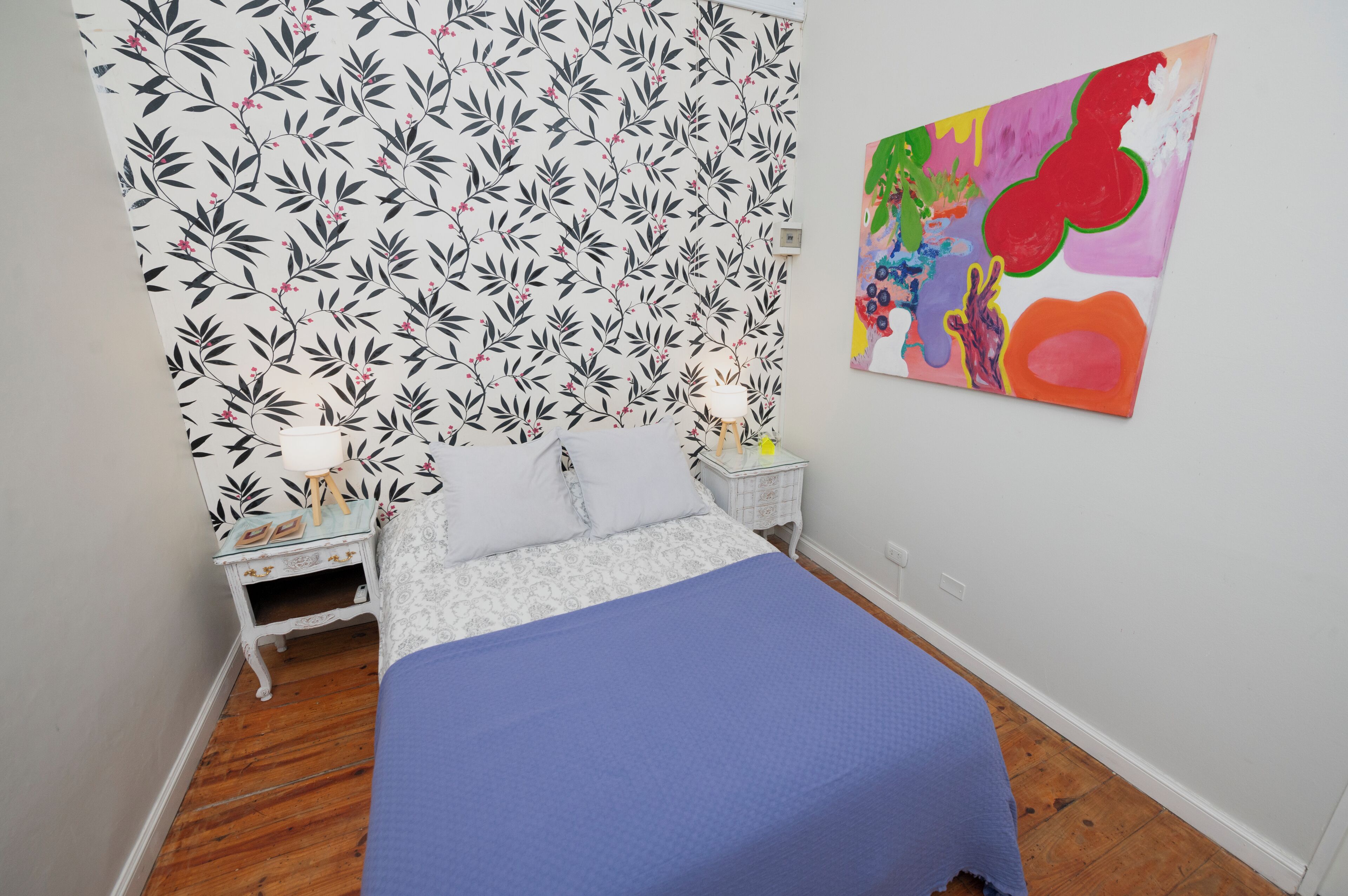 Une chambre avec un papier peint à motifs, un lit recouvert d’une courtepointe bleue, une table de chevet blanche avec une lampe, et un tableau coloré accroché au mur.