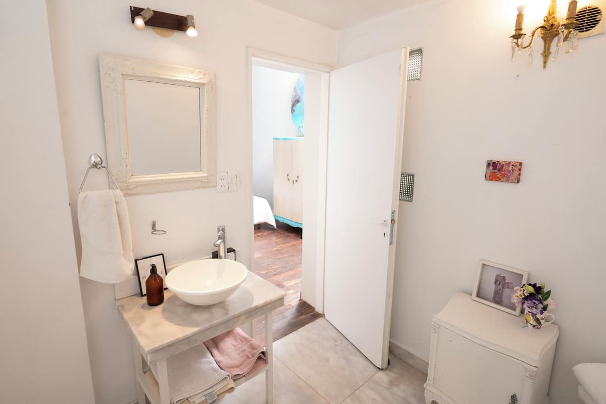 A bathroom with a white sink, a mirror, a towel rack, a small cabinet, and a shower area visible through an open door.