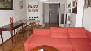 A living room with a red sofa, a wooden coffee table, a dining table with chairs, and a bookshelf.