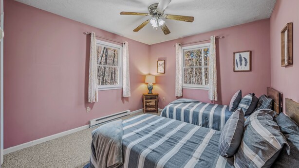A bedroom with two beds, a ceiling fan, a window with curtains, a bedside table with a lamp, and a framed picture on the wall. - Namba Cabin - Our Cabin (Great Cacapon)