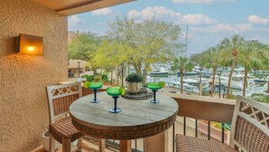 A balcony with a round table, wicker chairs, and a view of a marina with boats and palm trees. - 7104 Harbourside 1 @ Shelter Cove | Pool | Stunning Marina Views (Hilton Head Island)
