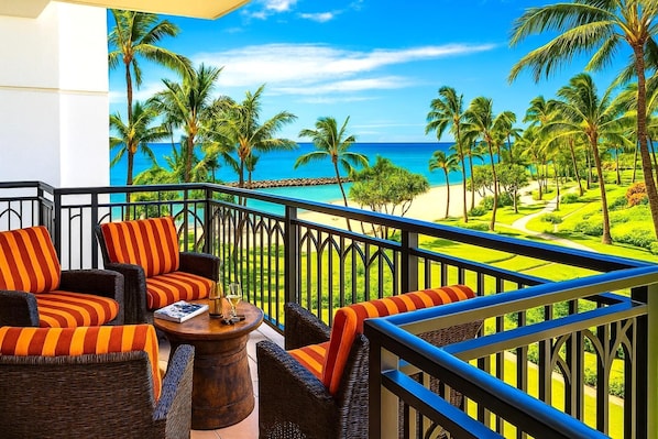 A balcony with wicker furniture and striped cushions, overlooking a beach with palm trees and the ocean.