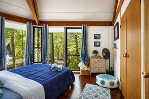 A bedroom with a large bed, a wooden wardrobe, a bedside table with a fan, and a view of trees through the window. - Sleep in a Treehouse!! (Lake Ariel)