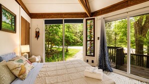 A bedroom with a large bed, a nightstand, a lamp, a window with a view of greenery, and a sliding glass door to an outdoor deck.