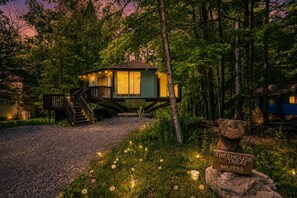 A treehouse with a wooden deck and a staircase leading up to it, surrounded by dense forest.