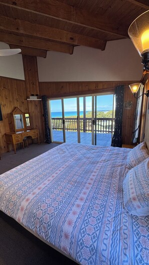 Une chambre avec un grand lit, des poutres apparentes en bois au plafond, une commode et une vue sur l’océan par une porte coulissante.