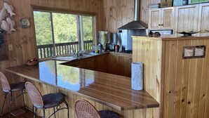 A wooden interior with a kitchen area, bar stools, and a large window. - Ocean View Holiday Home Orchid Beach | Phippsy's On Fraser | Sleeps 20 people! (K'gari)