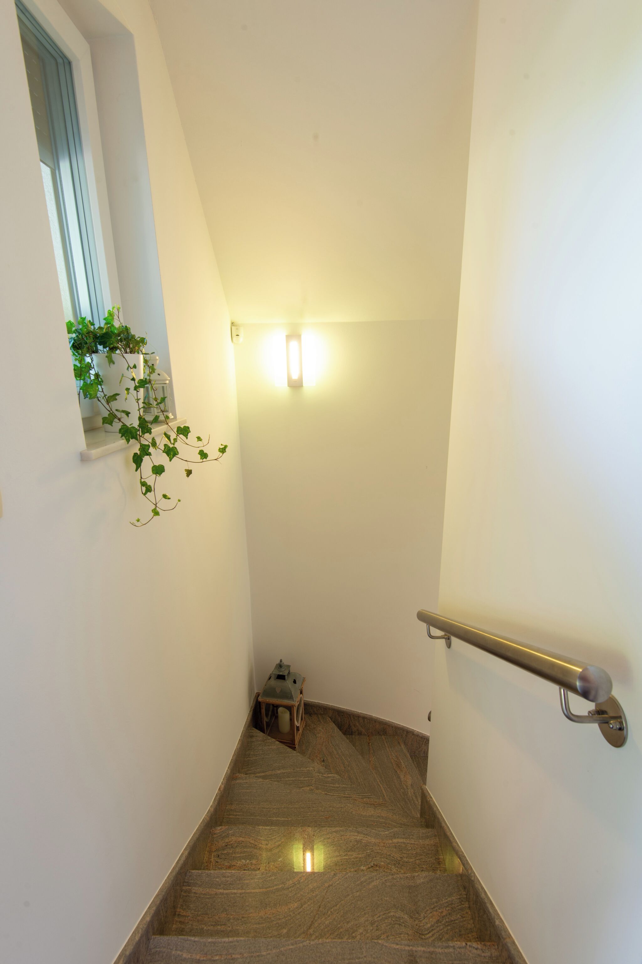 A narrow staircase with a handrail, a small window with a plant, and a light fixture at the top.