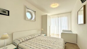 A bedroom with a bed, bedside table, lamp, window with curtains, and a mirror. - Villa Emy 11 Fronte Mare - Carraro Immobiliare Jesolo (Lido di Jesolo)