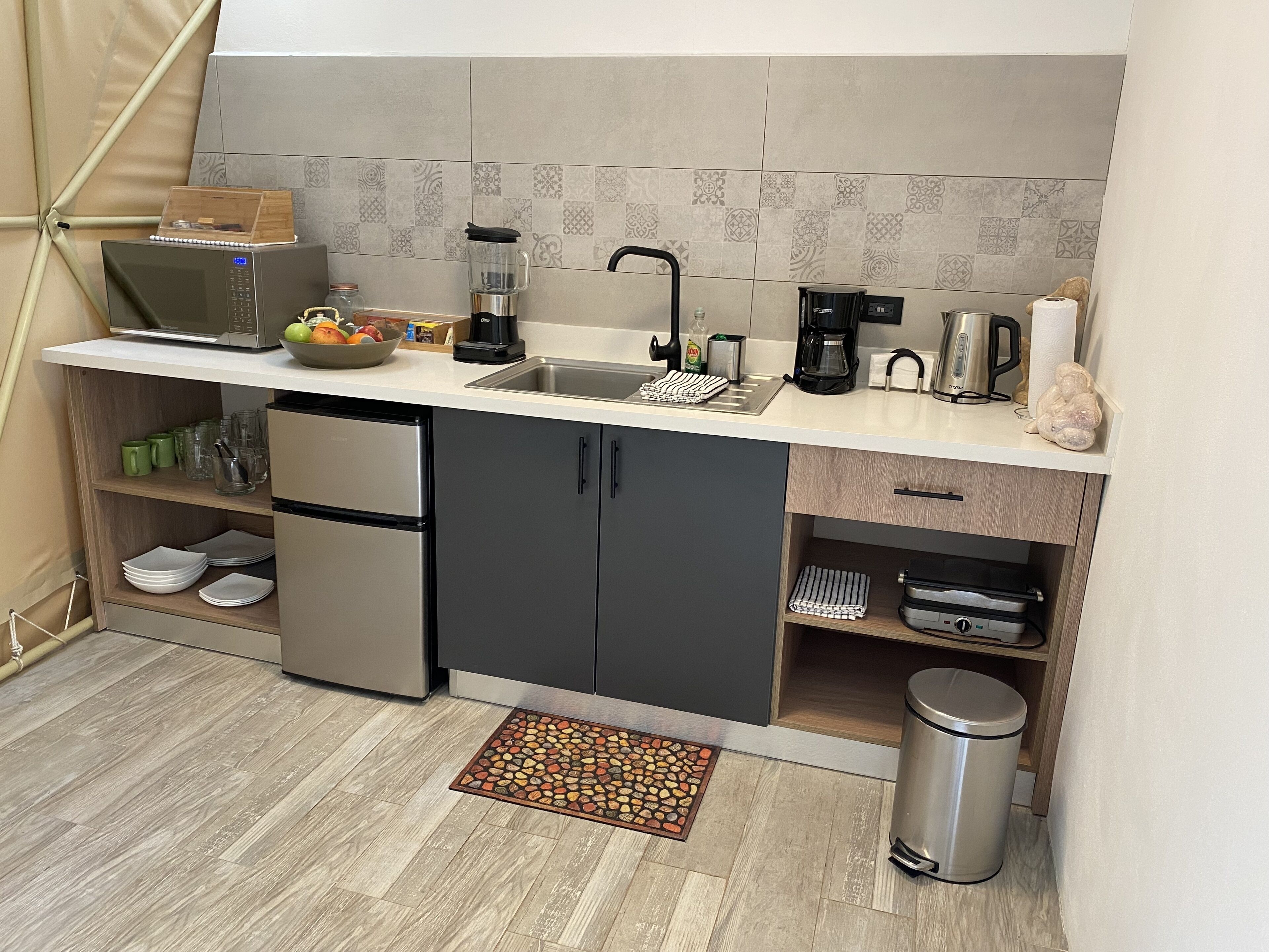 Cocina compacta con microondas, cafetera y lavabo. Se observan armarios, un cubo de basura y una pequeña alfombra.