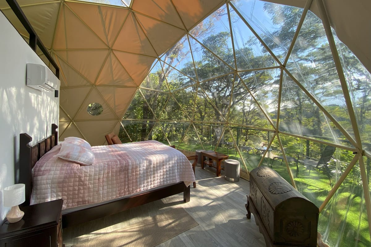 A bedroom with a bed, bedside table, and a view of the outdoors through a geodesic dome structure.