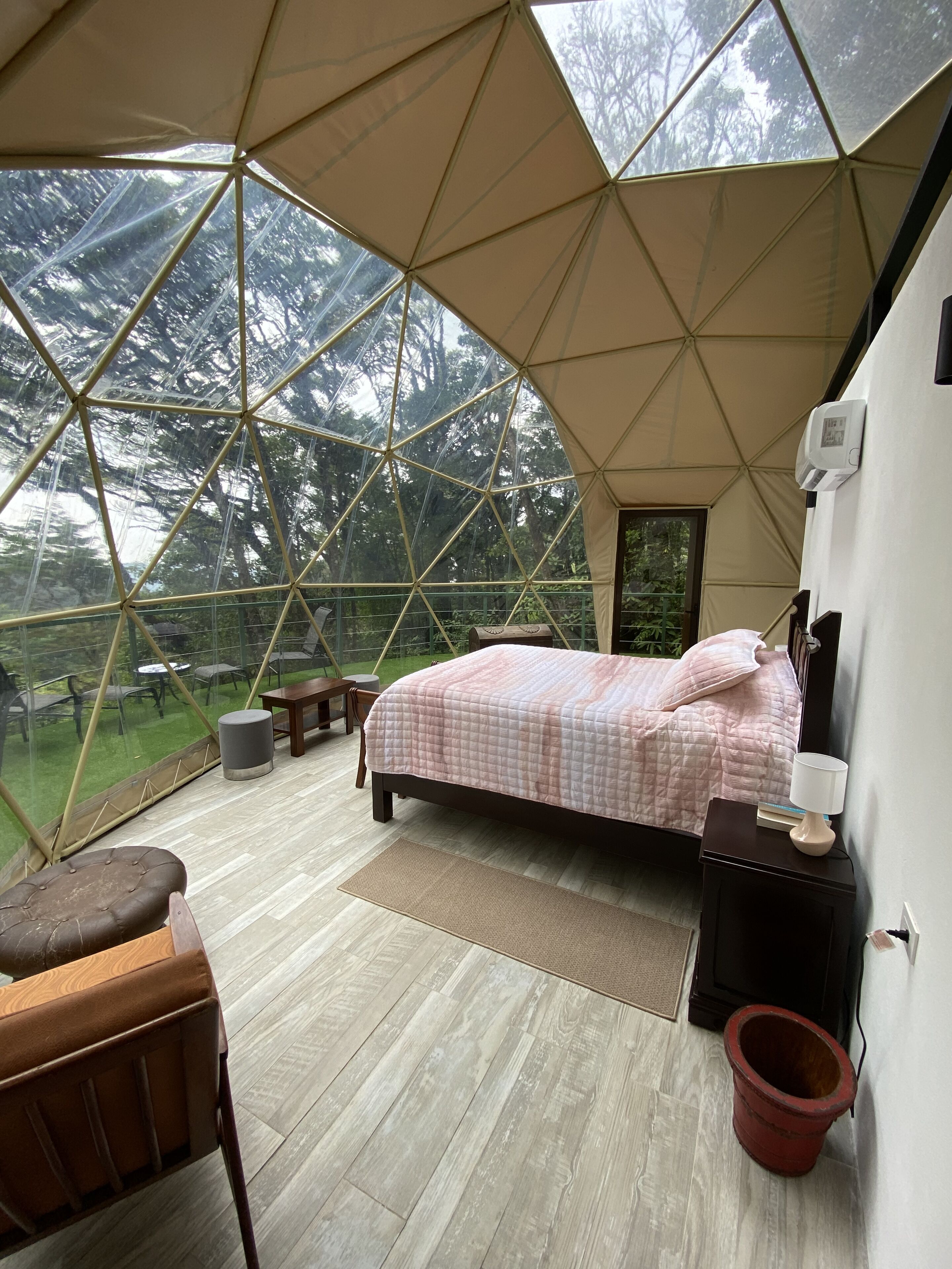 A bedroom with a bed, bedside table, lamp, and a view of the outdoors through a geodesic dome structure.