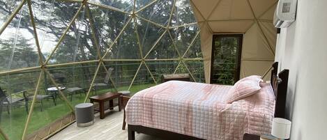 A bedroom with a bed, bedside table, lamp, and a view of the outdoors through a geodesic dome structure.