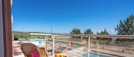 A balcony with a wicker chair, a bowl of fruit, and a view of a swimming pool and greenery.