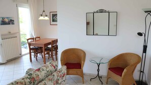 Un soggiorno con un divano, due poltrone in vimini con cuscini rossi, un tavolo da pranzo con sedie, un tavolino da caffè in vetro, una lampada da terra e uno specchio fissato alla parete.