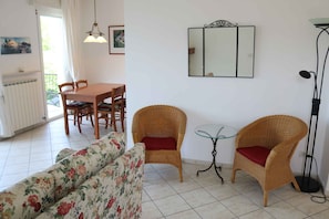 A living room with a sofa, two wicker chairs with red cushions, a dining table with chairs, a glass coffee table, a floor lamp, and a wall-mounted mirror.