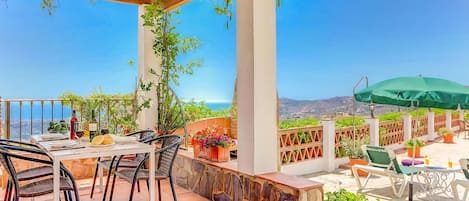 An outdoor patio with a table set for dining, surrounded by potted plants and a view of the sea.