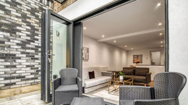 A modern living area with wicker furniture, a glass door leading to a balcony, and a brick wall. - Glamourised 1 Bed Apt with Private Garden (London)