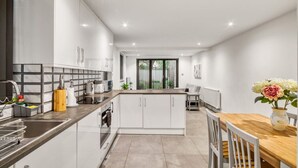 A modern kitchen with white cabinetry, a central island, and a dining area with a wooden table and chairs. - Glamourised 1 Bed Apt with Private Garden (London)