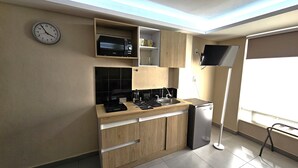 Una cucina compatta con forno a microonde, piano cottura e frigorifero, pensili in legno e una sveglia appesa al muro.