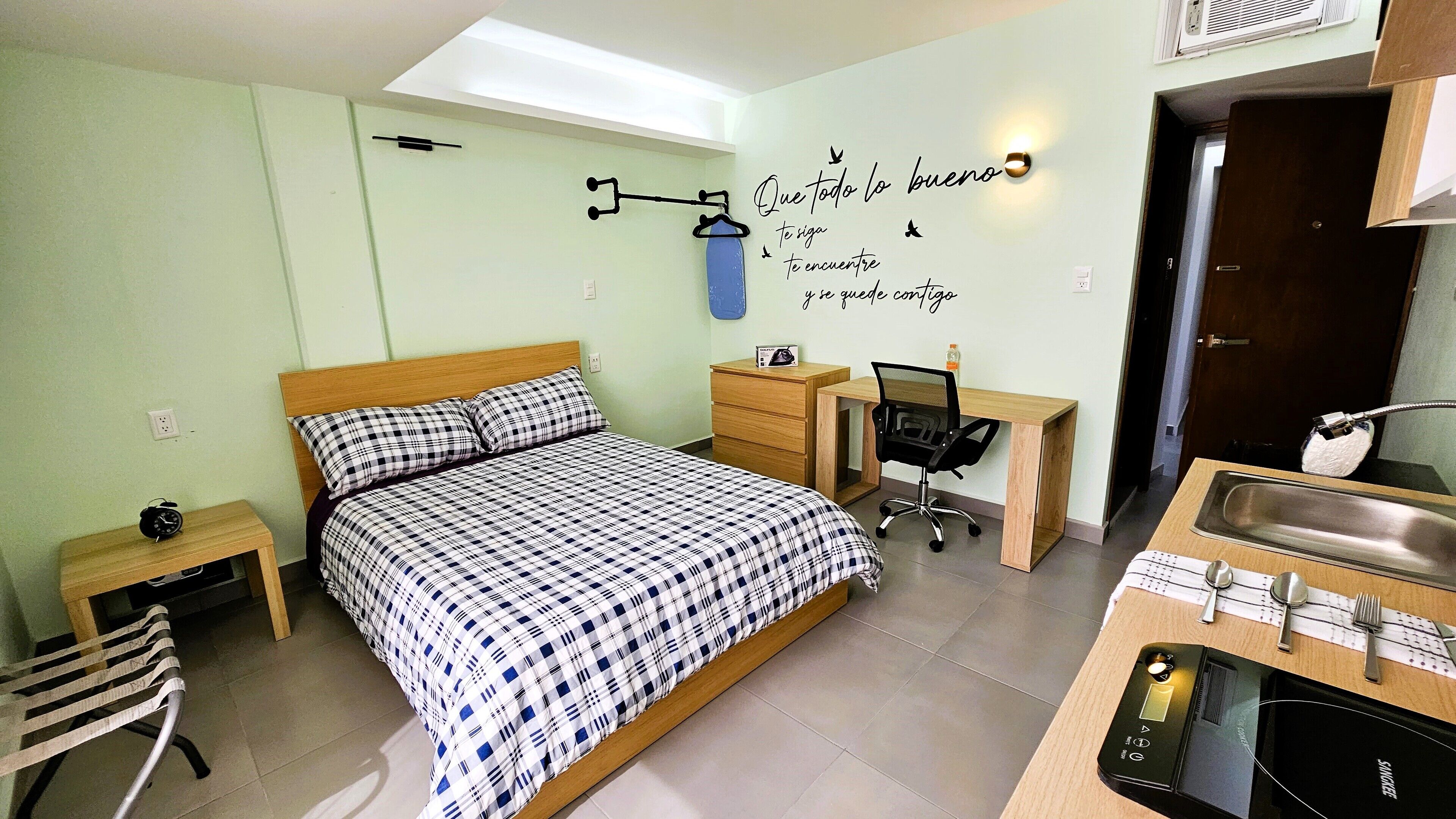 Un dormitorio con cama, escritorio, silla, mesita, perchero, reloj de pared y luz instalada en la pared.