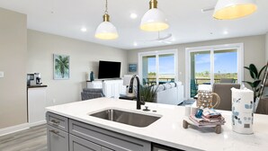 A modern kitchen with a large island, white countertops, and stainless steel appliances. There are pendant lights above the island and a ceiling fan.