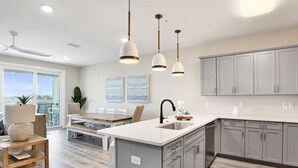 A modern kitchen with a large island, grey cabinets, pendant lighting, and a dining area with a view. - Serenity Sunrise: Gulf Shores Luxury Condo (Gulf Shores)