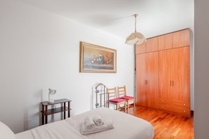 A bedroom with a bed, a bedside table with a lamp, a chair, a wooden wardrobe, and a painting on the wall.