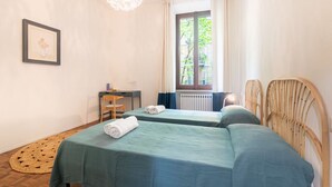 A room with two beds, a desk, a chair, a woven rug, and a window with curtains. - Comfortable apartment located on the second floor, of four in total, of a 1930/40s building with no elevator.In 75 square meters of space it comfortably accommodates four people with one bedroom with a double bed and one bedroom with two single beds.It i (Parma)