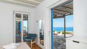 A bedroom with a bed, a chair, a wardrobe, and a view of the sea through an open door. - Private 1-Bedroom Villa Suite in Mandrakia with Swimming Pool (Mandrakia)