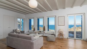 A spacious living room with a large grey sofa, wooden flooring, and large windows offering a view of the sea. - Private 1-Bedroom Villa Suite in Mandrakia with Swimming Pool (Mandrakia)