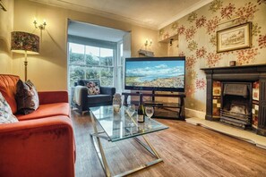 A living room with a sofa, a glass coffee table, a television, a fireplace, and a large window. - Sutherlands, Tenby - Six Bedroom Cottage, Sleeps 10 (Tenby)