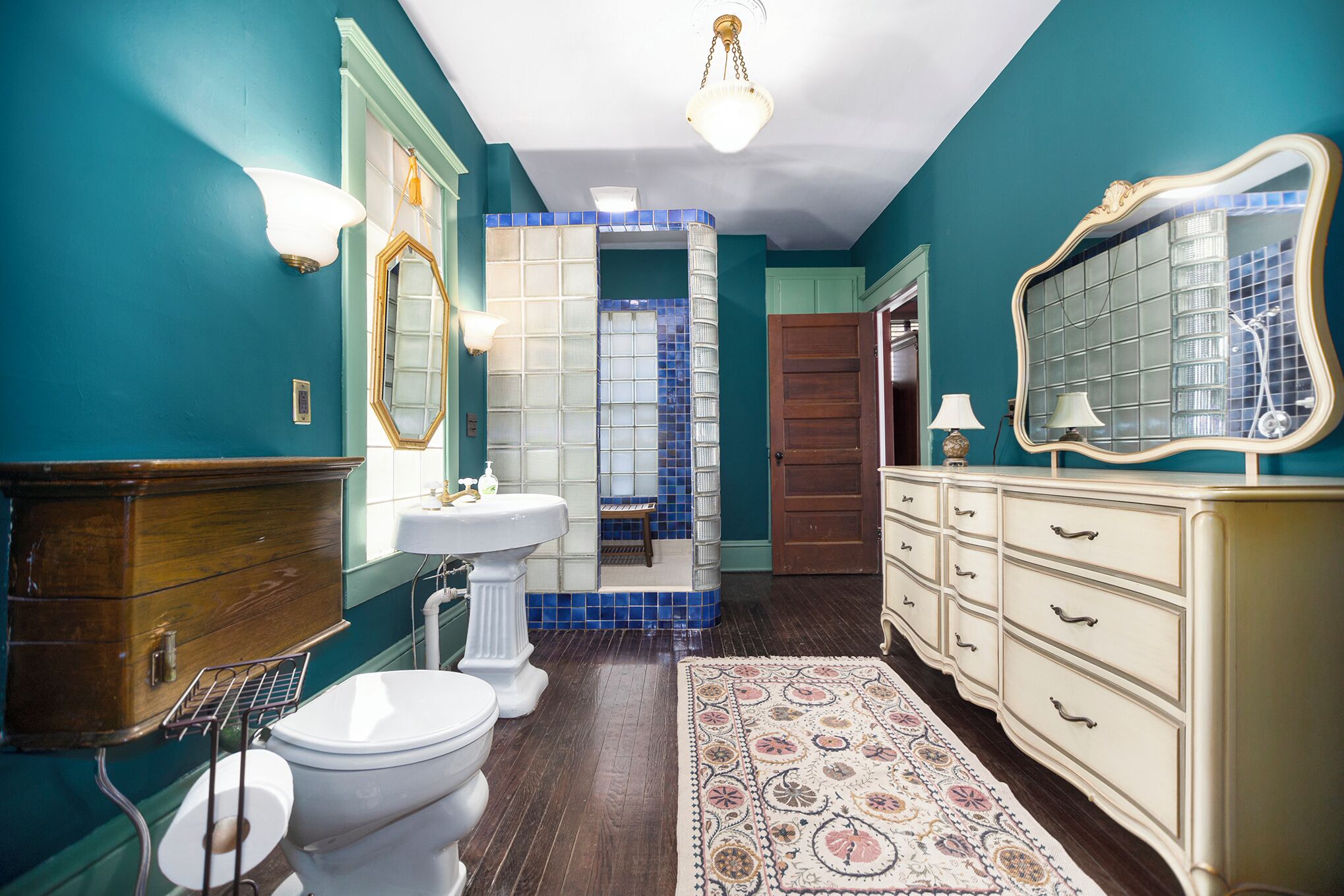 Un baño con paredes color turquesa, un lavabo blanco, un inodoro blanco, un mueble de madera y una alfombra estampada.