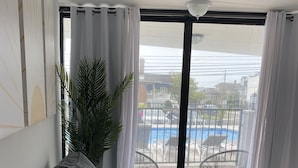 A modern living room with a grey sofa, a yellow decorative pillow, a potted plant, and a view of a swimming pool through large windows. - Poolside Condo - Just 2 blocks from the Beach, Boardwalk and Seaport Pier!  (North Wildwood)