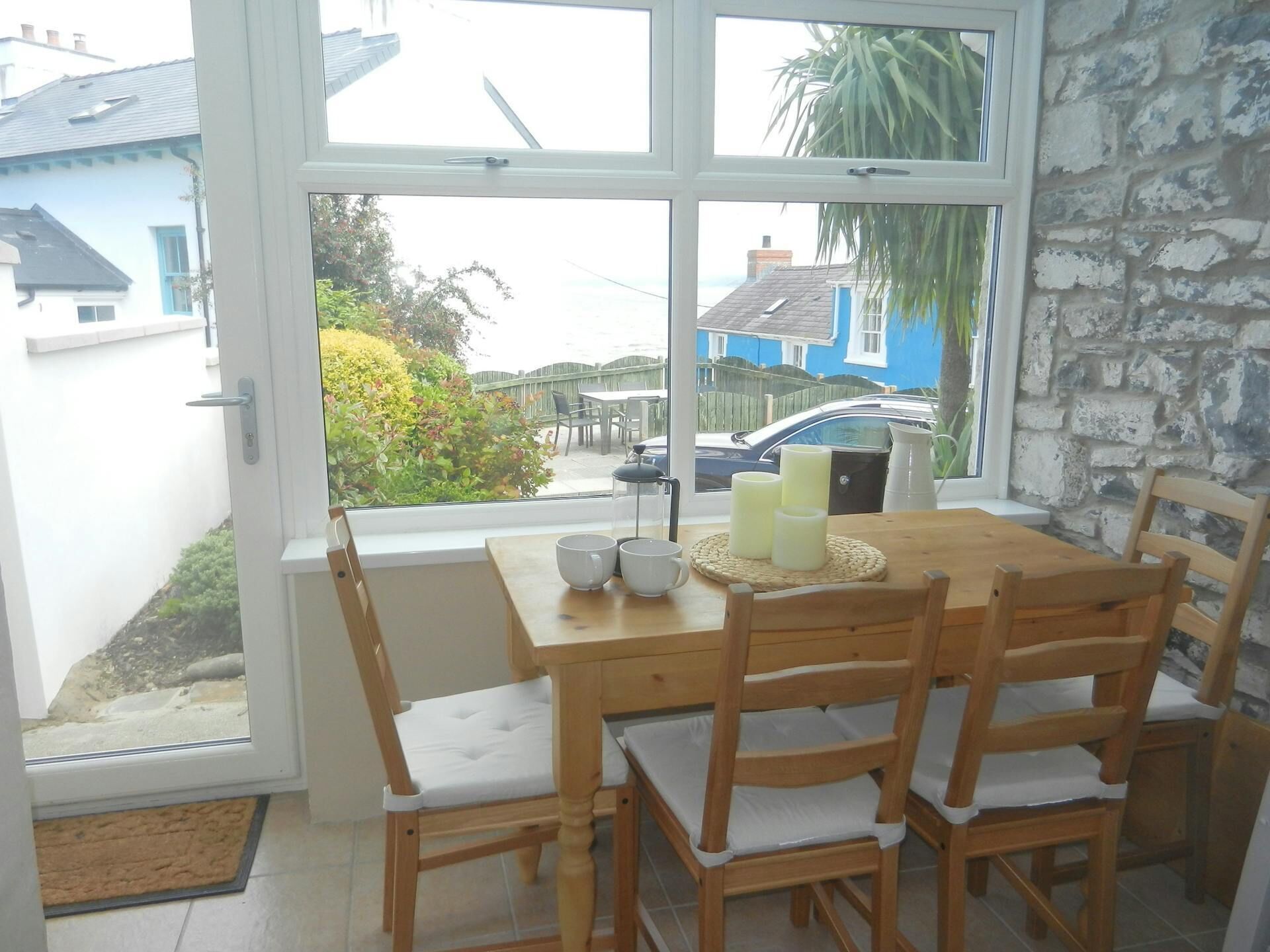 A dining area with a wooden table and chairs, a window with a view of houses and a car, and a mat at the door.