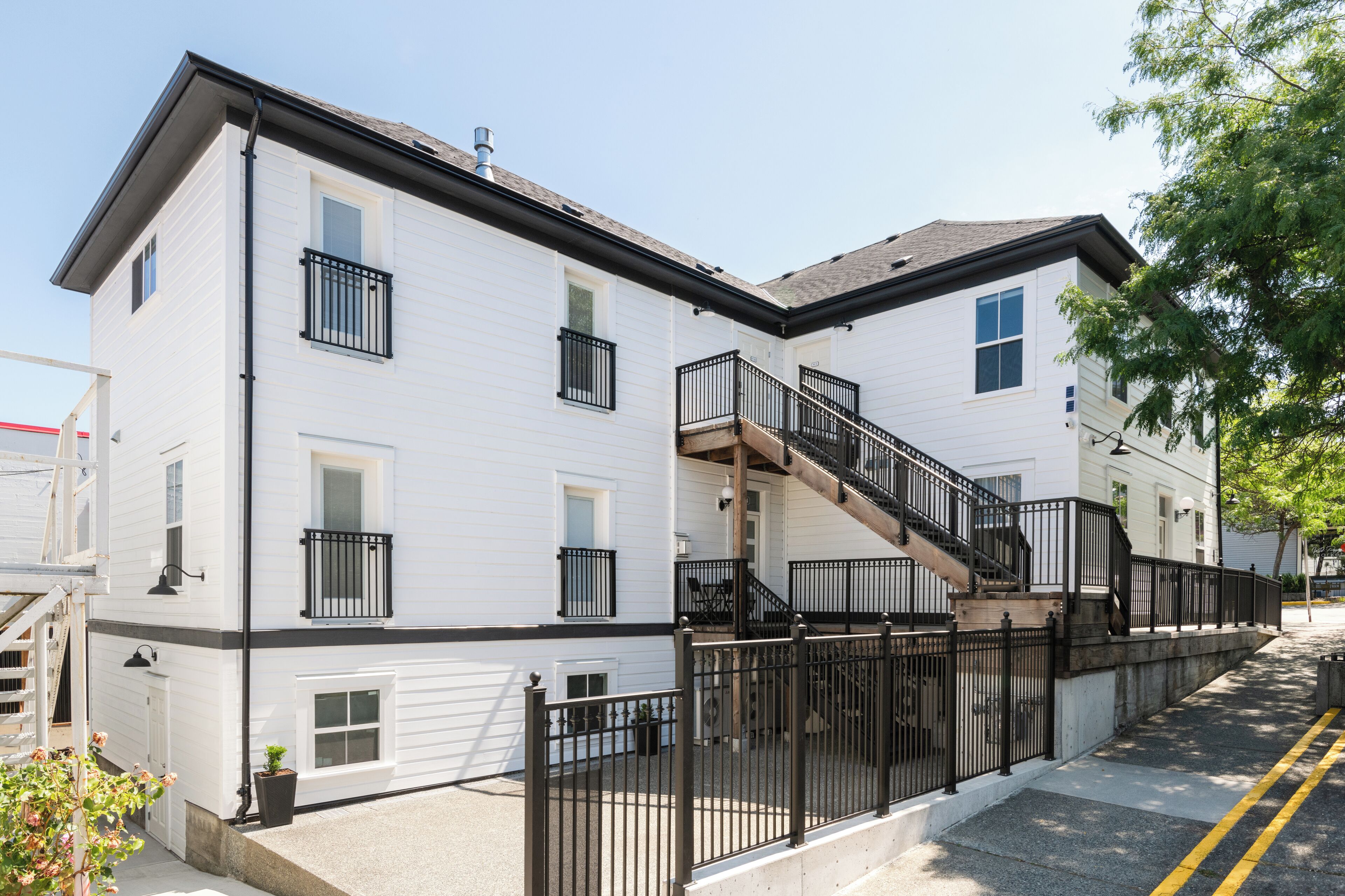 Un edificio residencial de dos pisos con una escalera y barandal de metal negro, revestimiento blanco y un camino de entrada con portón.