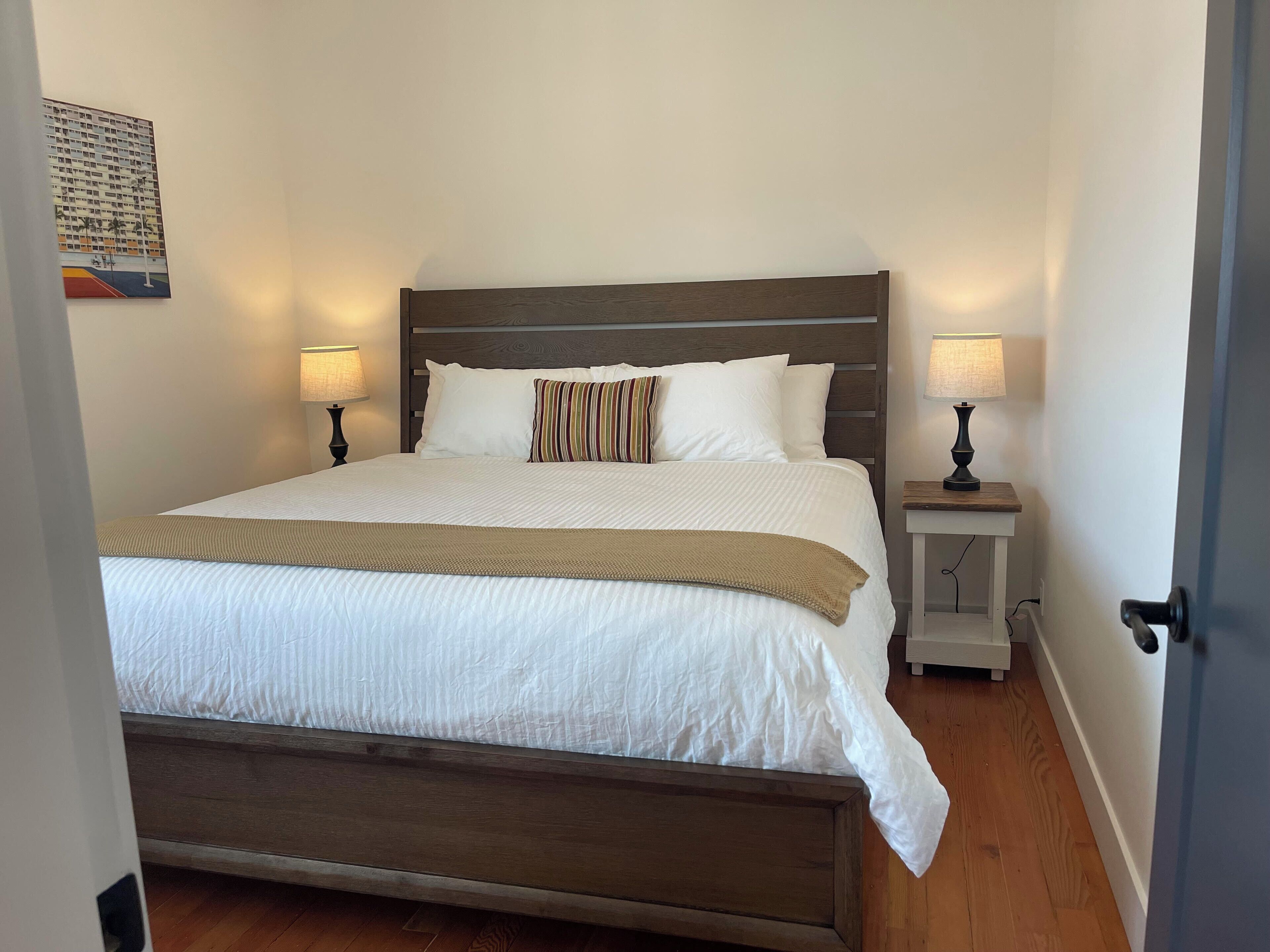 A neatly made bed with a wooden headboard, white bedding, and striped pillows. There are two bedside tables with lamps, and a small nightstand with a lamp. The room has wooden flooring and a plain wall with a framed picture.
