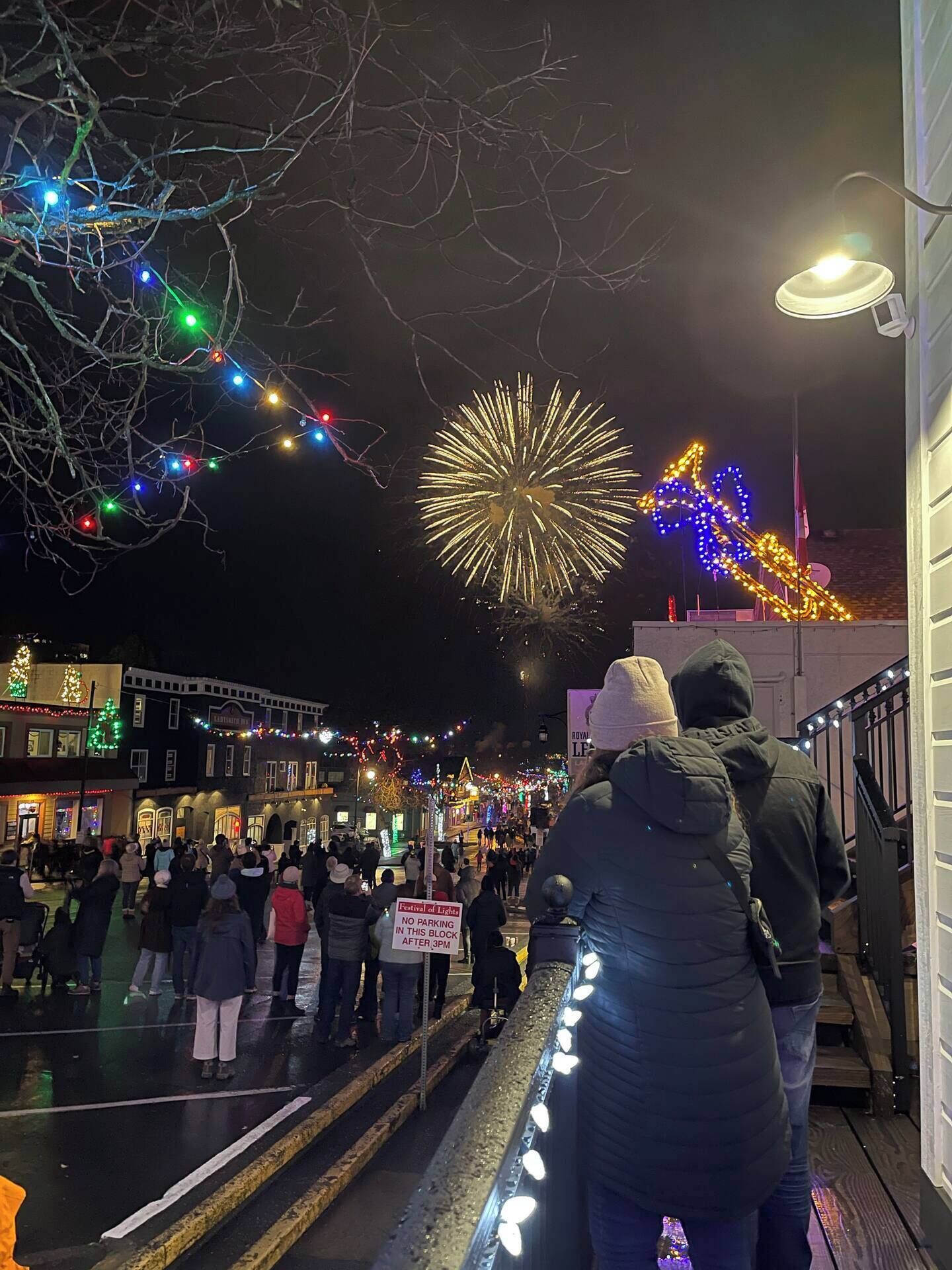 Une scène de rue nocturne, où une foule de personnes se trouve, illuminée par des lumières colorées et un grand feu d'artifice.