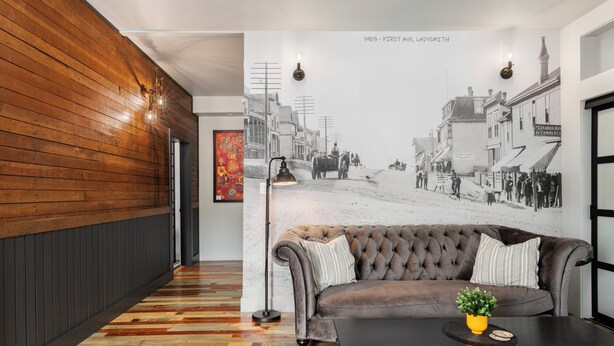 A hallway with a wooden wall, a black floor, a brown sofa with striped pillows, a black table with a yellow vase and a plant, and a large black-and-white street scene mural on the wall. - Temperance Hotel (Ladysmith)