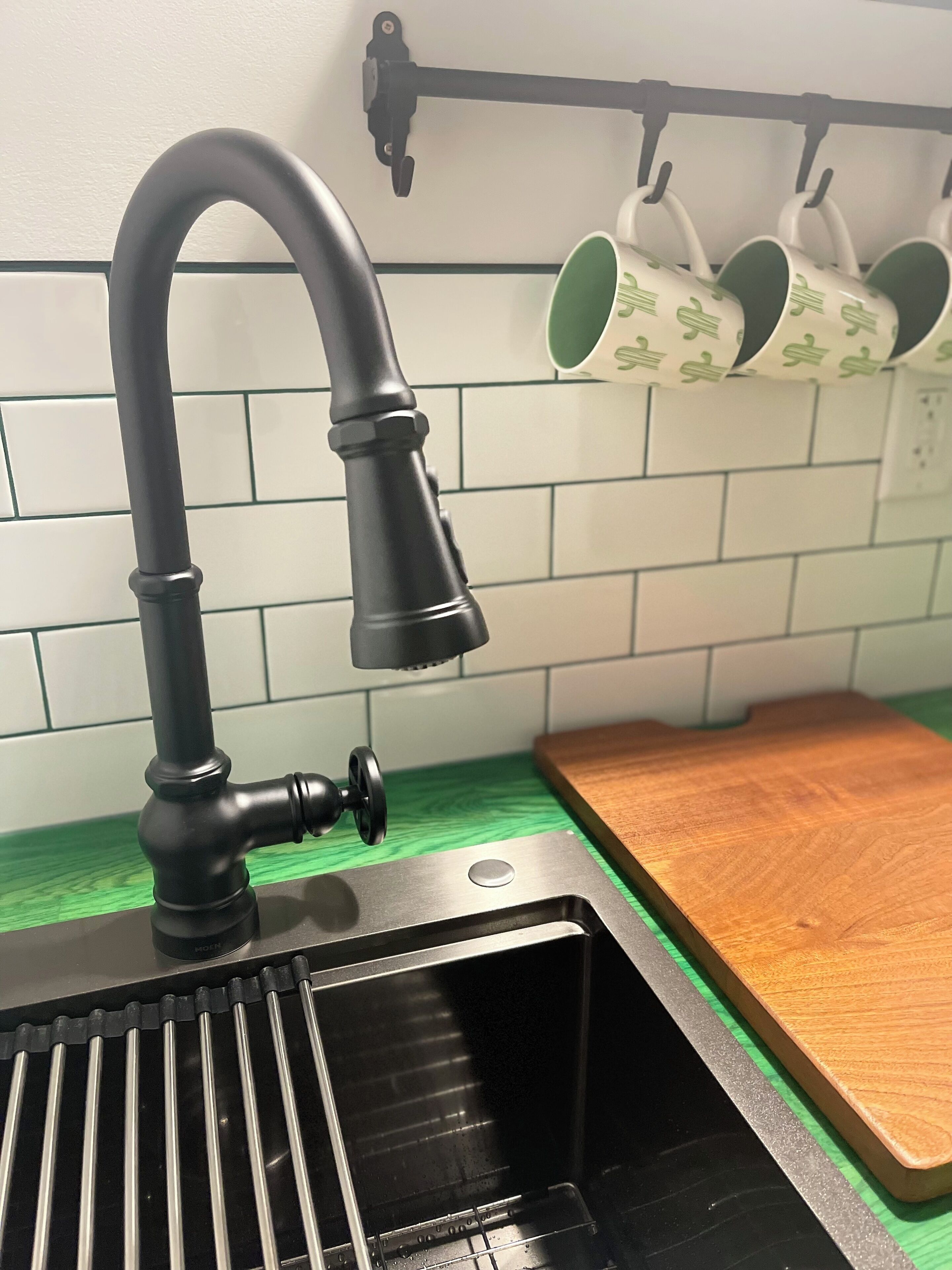 A kitchen with a black faucet, a wooden cutting board, and a rack holding green and white cups.
