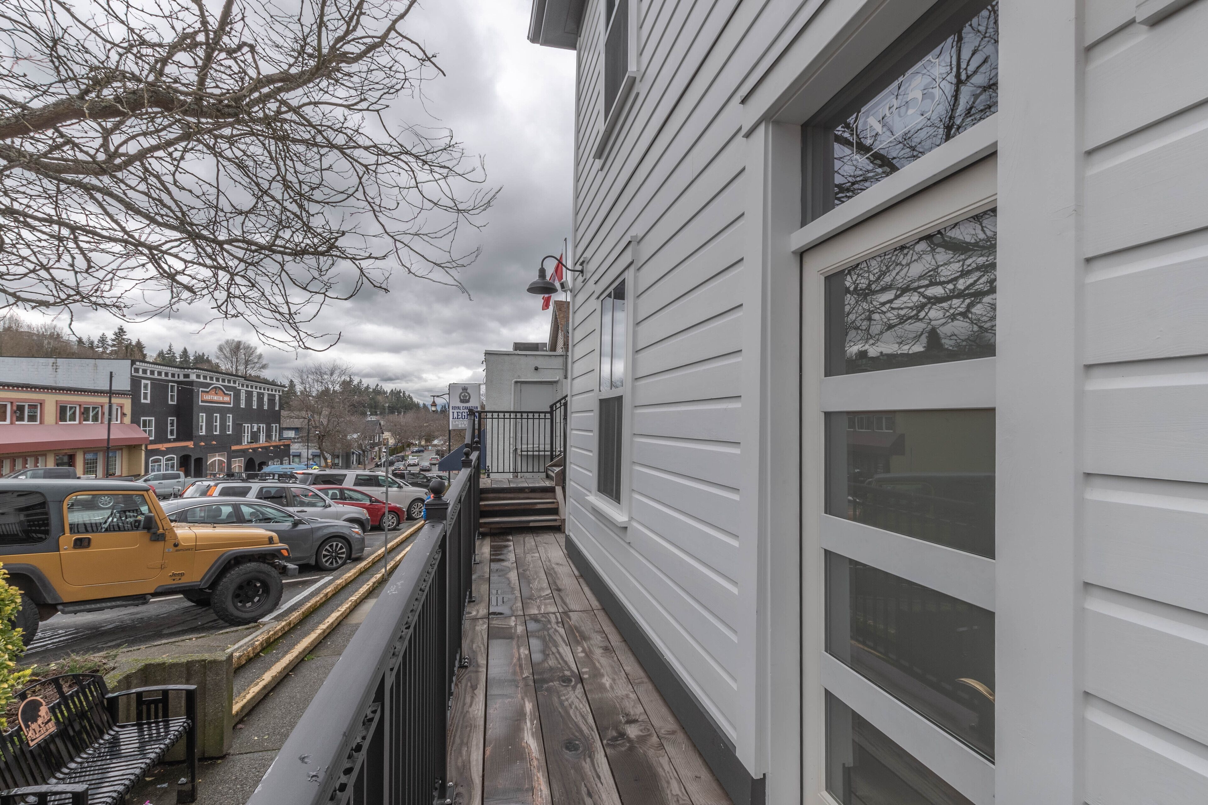 Une étroite terrasse en bois avec un banc, une Jeep jaune garée, et une rangée de voitures stationnées le long d’une rue, avec des bâtiments en arrière-plan.
