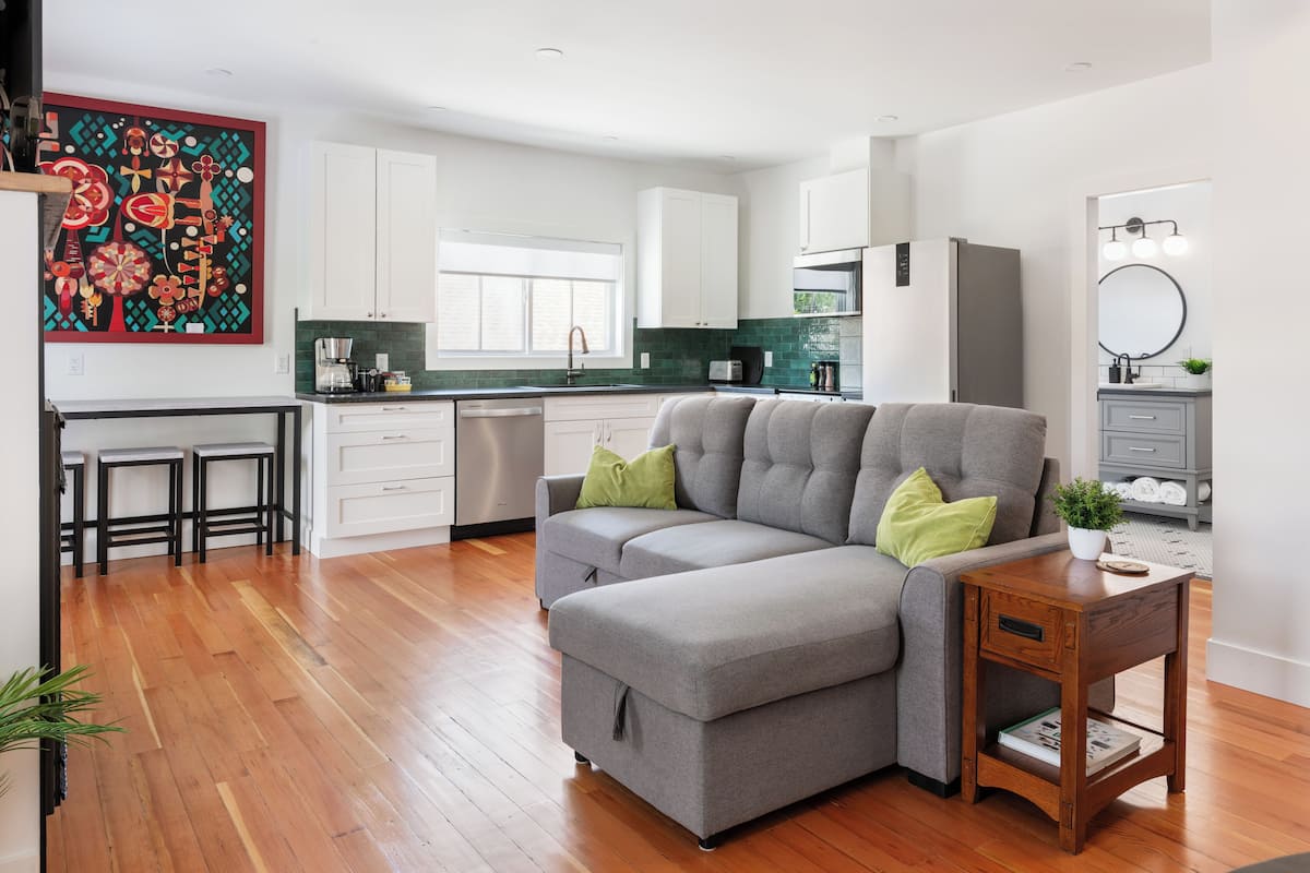 A modern kitchen with white cabinets, a grey sofa, and a wooden side table.