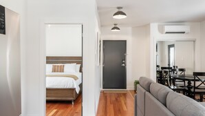 A modern hotel room with a wooden floor, a bed with white bedding, a gray sofa, a dining table with chairs, and an air conditioning unit. - Temperance Hotel (Ladysmith)